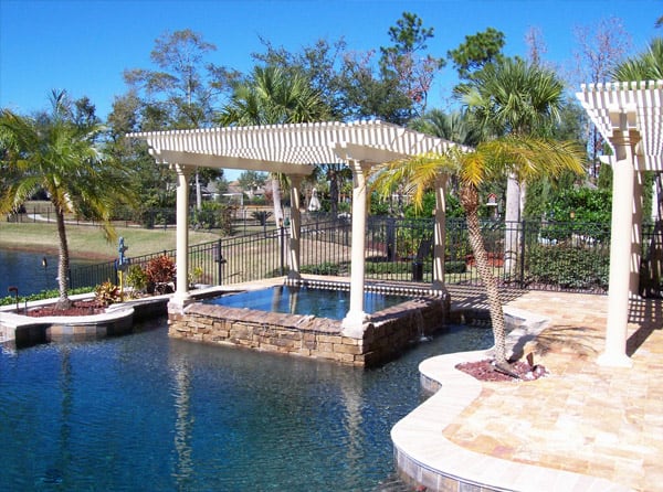 A white aluminum pergola in a backyard on top of a pool with trees in the background