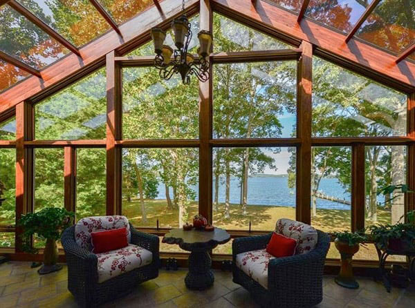 A cathedral sunroom with two comfortable chairs and a table next to glass walls overlooking a lake. 